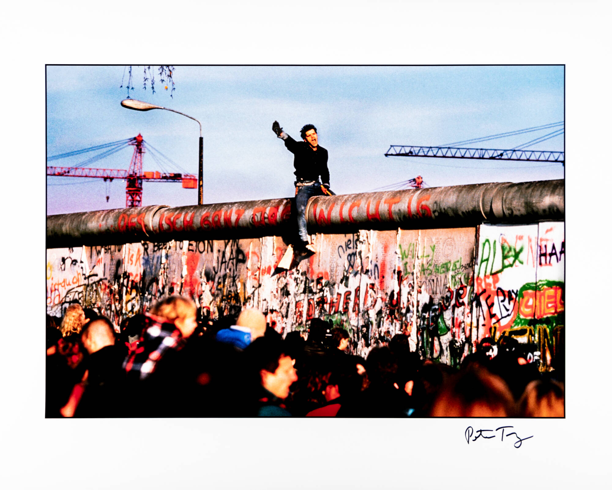 Photograph of a man sitting atop the Berlin Wall. The wall is covered in text most of which is in German. There is a crowd of people standing around the wall below the man.